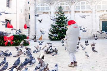 Wall Mural - Cute child girl is feeding doves on square in Europe. Kid is scared of flying pigeons. Christmas romantic vintage atmosphere in old town. Authentic, ancient architecture in Dubrovnik, Croatia