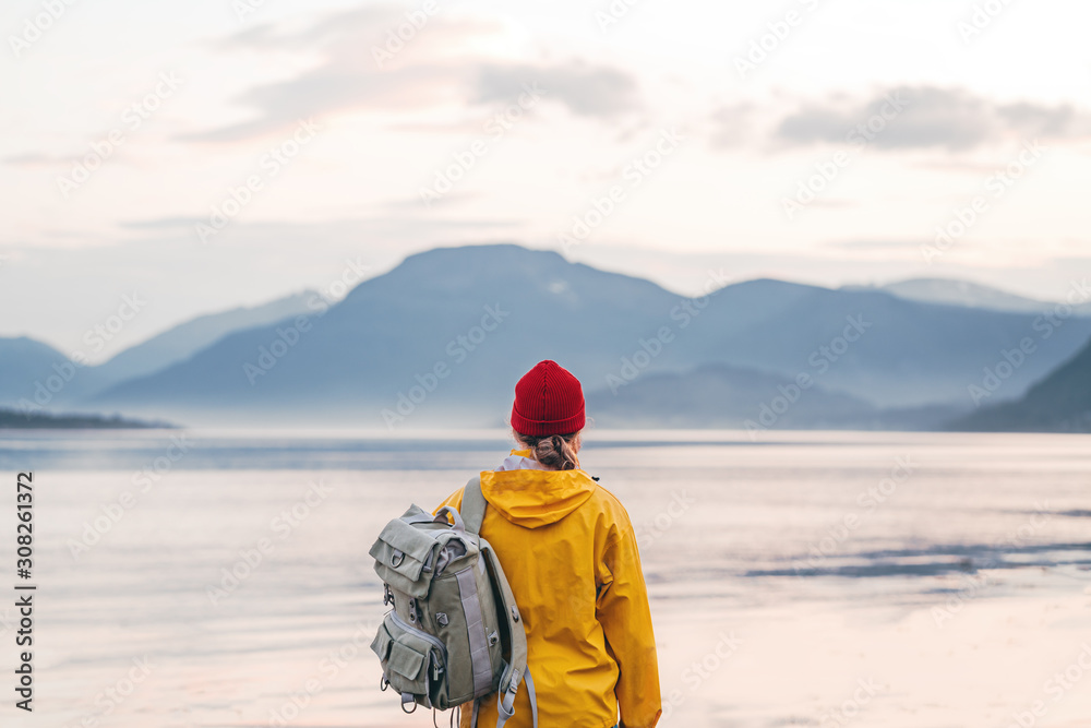 Alone traveler wearing yellow raincoat and backpack looking at fantastic fjord and mountain landscape. Lifestyle outdoor adventure, scandinavian wanderlust - obrazy, fototapety, plakaty 