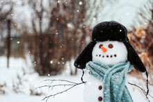  Snowman In A Blue Knitted Scarf And Hat Under A Snowfall Copy Space.