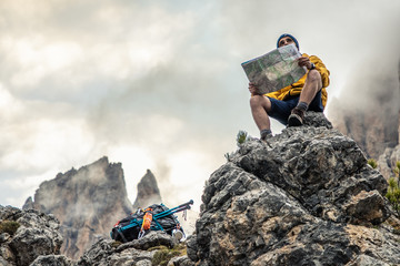 Wall Mural - young man hiker sitting on stone mountain reading map, with cloudy sky and fog. yellow jacket, backp