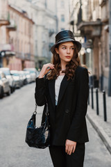 Outdoor fashion portrait of young elegant confident model, woman wearing trendy faux leather bucket hat, black suit, holding stylish hobo bag, handbag, posing in street of European city