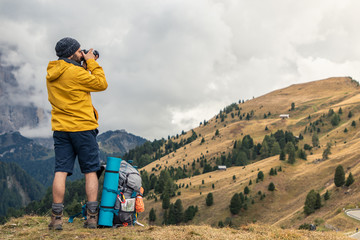 Wall Mural - young man hiker taking pictures on mountain hill with cloudy sky. yellow jacket, backpack, black bea