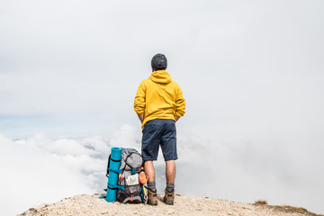 Wall Mural - young man hiker standing on mountain peak with cloudy sky and fog. yellow jacket, backpack and beani
