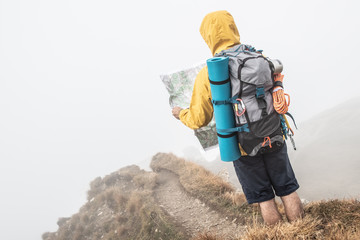 Wall Mural - young man hiker on mountain trail reading map. cloudy sky and fog. yellow jacket, backpack and beani