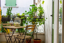 An Italian Balcony With Green Potted Plants And Garden Furniture. A Table And Chairs To Enjoy The Balmy Evening.