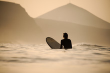 Rear View Woman In The Black Diving Suit Sitting On The Surfing Board In The Sunset Time