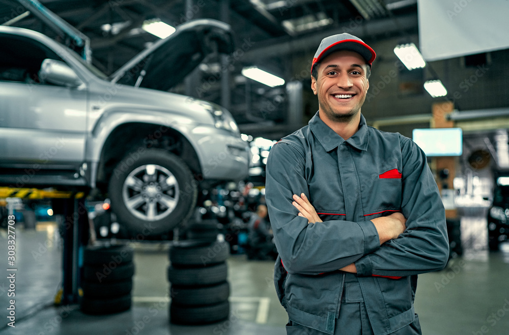 Handsome auto service mechanic in uniform is standing on the background of car with open hood, smiling and looking at camera. Car repair and maintenance. - obrazy, fototapety, plakaty 
