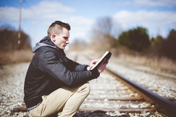 Sticker - Shallow focus shot of a male sitting down on train tracks while reading the bible