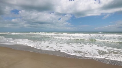 Wall Mural - ocean waves break on a sandy beach under a cloudy blue sky over a calm sea in this seamlessly loopin