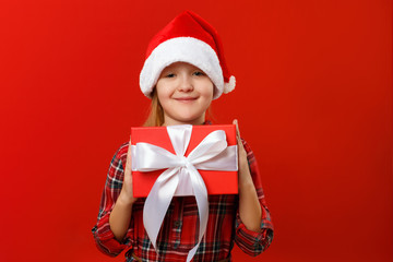 Wall Mural - Cute happy child holds a box with a gift. Charming little girl in santa hat and dress on a red background. Christmas and New Year concept