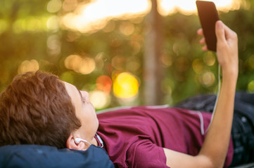 Teen boy with smartphone lies on the bench in the park.   Teenage boy is using phone, outdoors.  Caucasian teenager in casual clothes with cell phone in park. Soft focus effect.