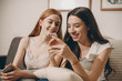 © Strelciuc - Two lovely caucasian girlfriend laughing while looking to a smartphone screen sitting on a sofa at home.