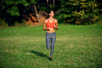 Wall Mural - Full length of attractive fit caucasian brunette in sportswear and with ponytail running on meadow. Sunny morning in nature.