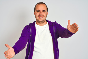 Sticker - Young man wearing purple sweatshirt standing over isolated white background looking at the camera smiling with open arms for hug. Cheerful expression embracing happiness.