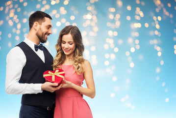 valentines day and people concept - happy couple with gist box in shape of heart over holiday lights on blue background