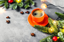 Christmas And New Year Composition With Orange Cup Of Coffee Surrounded By Fir Brunches With Red Balls And Lights On Gray Table.