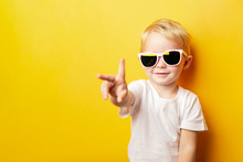 Portrait Of Cheerful Little Boy In A White T-shirt Wearing Sunglasses And Looking Away On Orange Background Showing Two Fingers Peace Sign