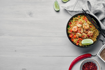 Wall Mural - Delicious fried rice with shrimps, lime and vegetables in a black bowl. Traditional Asian food on a white wooden table with a fabric tablecloth. Top view, directly above shot with space for text.