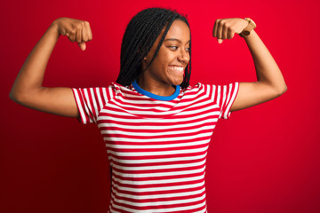 Canvas Print - Young african american woman wearing striped t-shirt standing over isolated red background showing arms muscles smiling proud. Fitness concept.