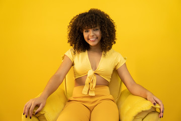 Wall Mural - Happy laughing American African woman with her curly hair on yellow background. Laughing curly woman in sweater touching her hair and looking at the camera.