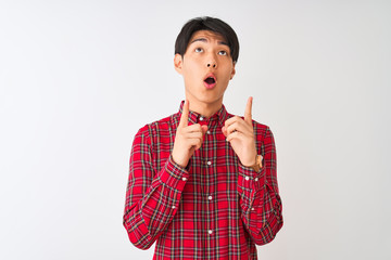 Wall Mural - Young chinese man wearing casual red shirt standing over isolated white background amazed and surprised looking up and pointing with fingers and raised arms.