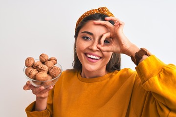 Poster - Young beautiful woman holding bowl with walnuts standing over isolated white background with happy face smiling doing ok sign with hand on eye looking through fingers