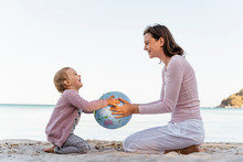 Happy Little Girl Sitting With Her Mother On The Beach Playing With Earth Beach Ball