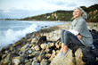 © Cavan Images - Side view of thoughtful senior woman sitting on rock at Manhattan Beach against sky during sunset