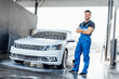 Professional washer in blue uniform washing luxury car stands near  car in bubbles.