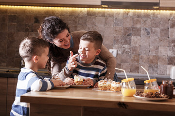 Mother  and her two little sons eating cakes at the kitchen.laughing and making fun.