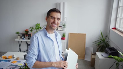 Wall Mural - Tilt up of papers and documents on desk and young entrepreneur standing in office, looking at app interface patterns on laptop screen, then closing pc and looking at camera with toothy smile on face