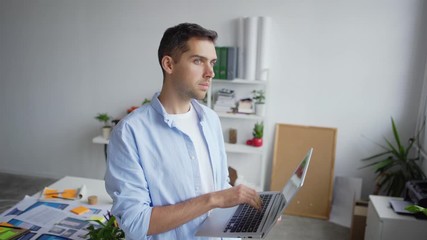 Wall Mural - Tilt up of papers and documents on desk and young entrepreneur standing in office with open laptop in his hands, then closing pc and looking away thoughtfully