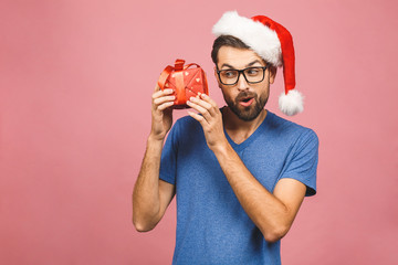 Wall Mural - Image of happy young man wearing christmas santa hat standing isolated over pink wall holding gifts box.