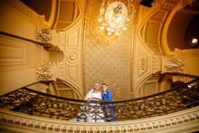 Beautiful Wedding Couple Stands Under A Golden Ceiling And Chic Chandelier In Opera. Indoors.