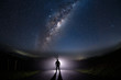 A mysterious man standing in the middle of the road looking into bright light with milky way starry night sky.