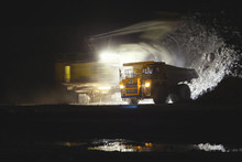 Blurry Image Of The Excavator In Motion On A Long Exposure Loading Stone Ore Into The Body Of A Mining Dump Truck, On A Dark Background With Artificial Illumination At Night.