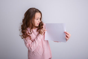 beautiful little brunette girl with a white sheet of paper on a white background, smiling, surprised