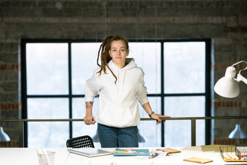 Pretty young designer in casualwear looking at you while standing by workplace