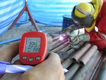 Tecnician Is Measuring The Temperature Of The Workpiece With A Temperature Gun. Focus The Human's Hand Holding A Infrared Thermometer Or Temperature Gun And The Background Is The Welder Who Is Welding