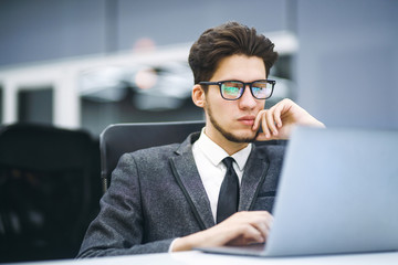 Business manager in glasses works on his laptop in the modern office. Young employee looking at computer monitor during working day in office. Young freelancer working from a laptop computer. 