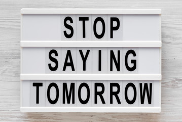 'Stop saying tomorrow' words on a lightbox on a white wooden background, top view. Overhead, from above, flat lay. Close-up.