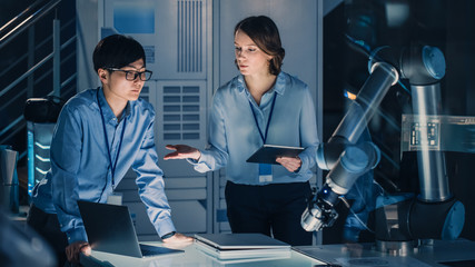 Fototapete - Man and Woman Engineers with Laptop and a Tablet Analyse and Discuss How a Futuristic Robotic Arm Works and Moves a Metal Object. They are in a High Tech Research Laboratory with Modern Equipment.