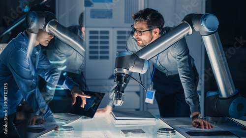 Diverse Team of Engineers with Laptop and a Tablet Analyse and Discuss How a Futuristic Robotic Arm Works and Moves a Metal Object. They are in a High Tech Research Laboratory with Modern Equipment.