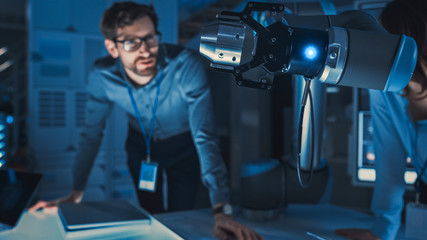 Poster - Close Up of a Futuristic Robotic Arm Picking Up a Metal Object and Moving It. Team of Engineers Following This Advanced Process. They are in a High Tech Research Laboratory with Modern Equipment.