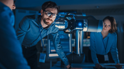 Poster - Futuristic Metal Robotic Arm Picking Up a Metal Object and Moving It. Senior Engineer Talks Through This Advanced Process. They are in a High Tech Research Laboratory with Modern Equipment.