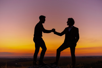 Silhouette of two men in sunset sky (orange and pink sky) on rock hill. Two gentlemen are taking, looking and agree each other, then shake hands. Agreement and cooperation concept