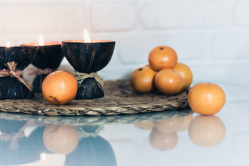 Wall Mural - Burning spa aroma candles in coconut shell, tangerines on wicker stand on a glass white table, cozy home interior background top view.