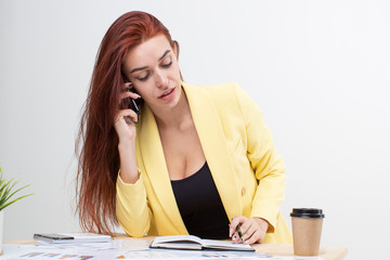 Wall Mural - A red-haired girl in a yellow business suit sits at a table and talks on the phone. Working day in the office