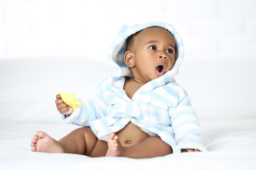 Wall Mural - American baby girl in bathrobe sitting on bed and holding toy