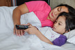 Asian Little Boy On Bed Using Remote Control To Watch Television while His Mother Asleep